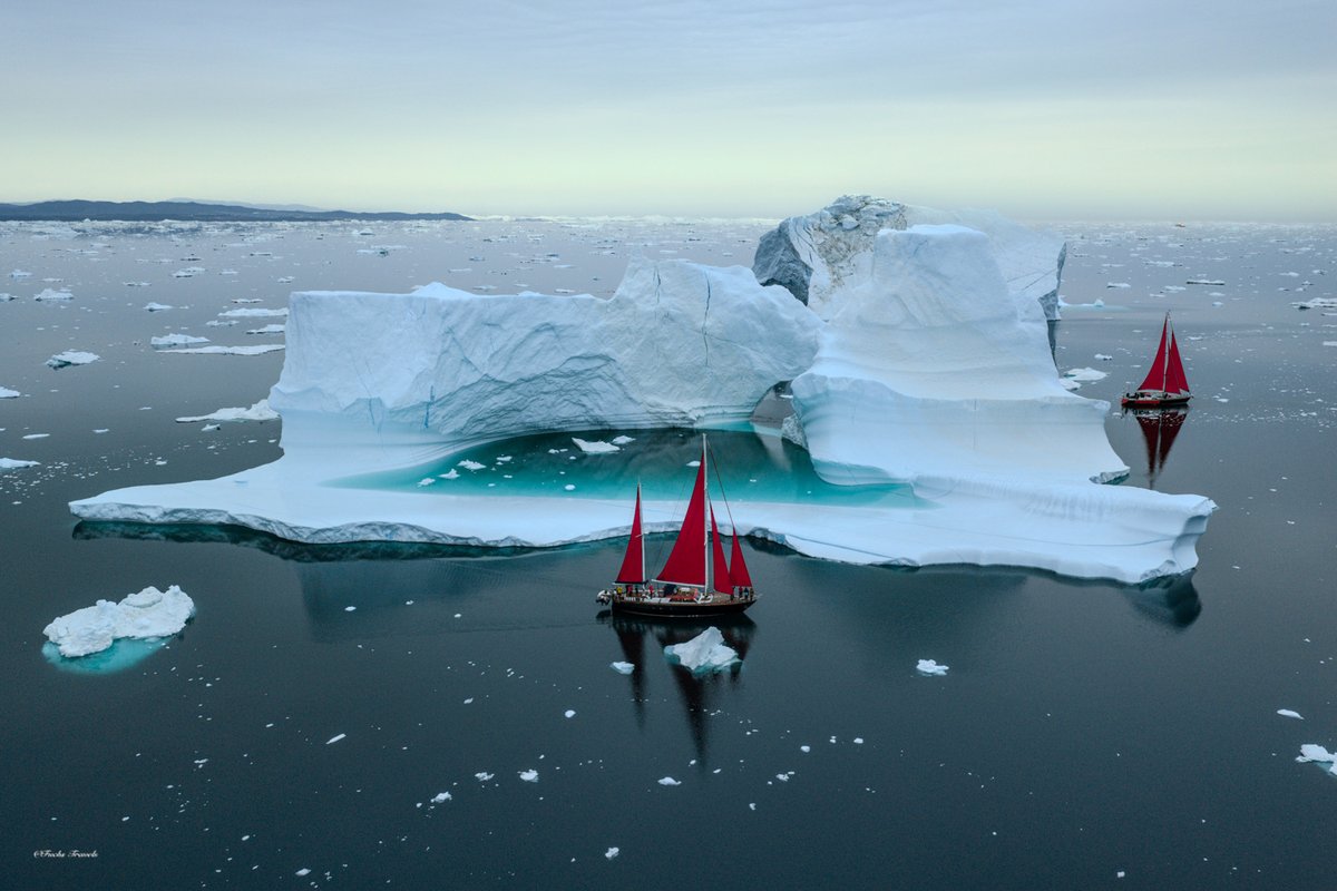Aerial drone photograph of two red-sailed schooners navigating among massive icebergs in turquoise Disko Bay waters off Ilulissat Greenland