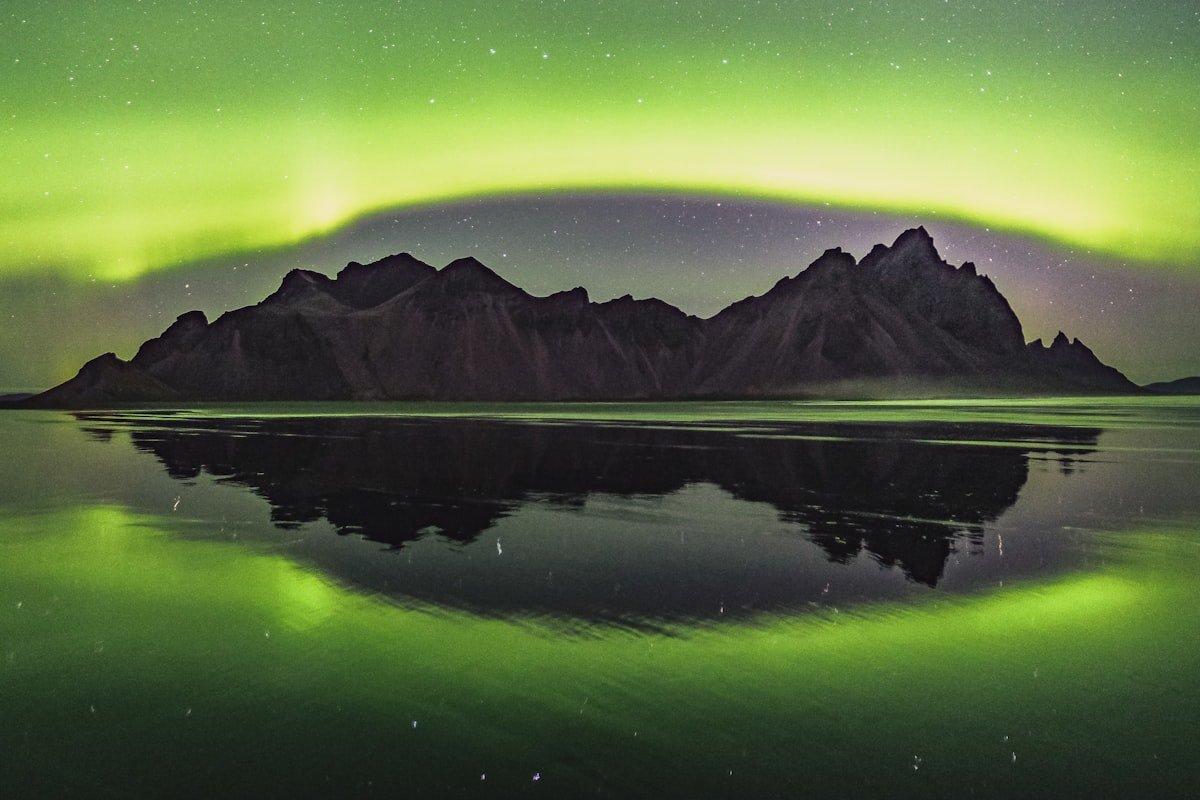 Vivid green northern lights arching over dramatic mountain peaks with perfect reflection in arctic waters