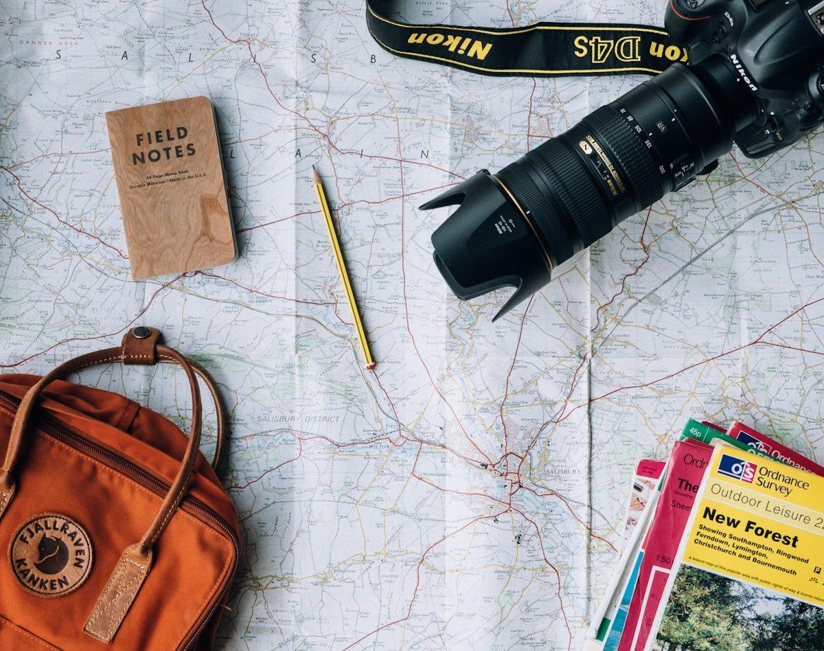 Flat lay of expedition planning essentials including camera with telephoto lens topographic map field notebook and travel guides
