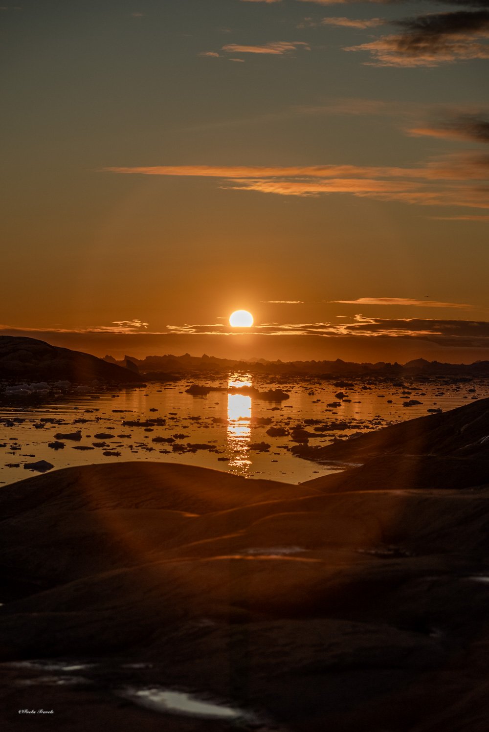 The midnight sun hanging low over Disko Bay with icebergs silhouetted along the horizon in warm golden light