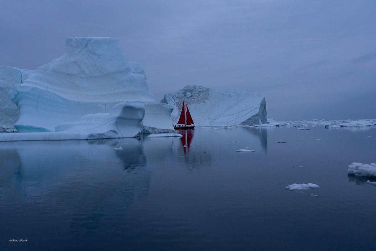 Red sailboat navigating between massive blue-white icebergs in moody atmospheric conditions