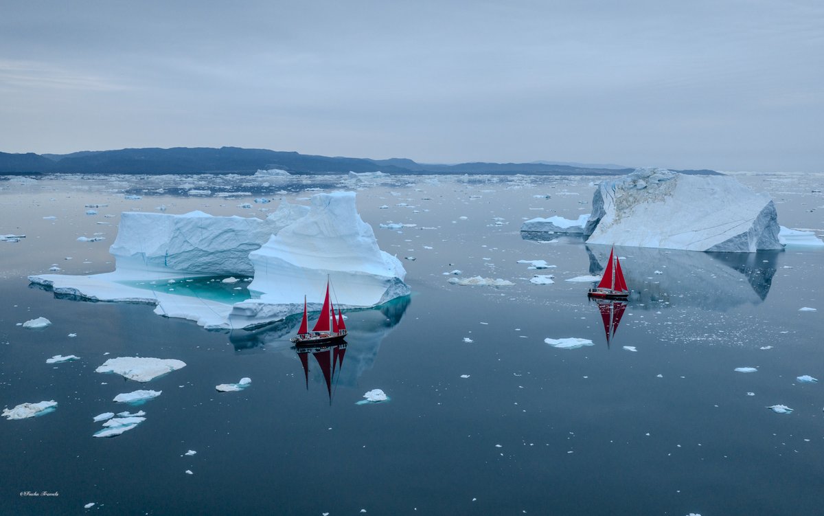 Wide aerial view of two red sailboats dwarfed by massive icebergs scattered across the vast expanse of Disko Bay