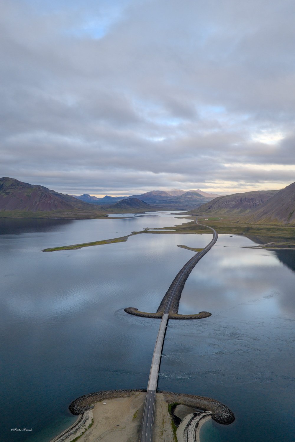 Winding causeway bridge crossing a serene Icelandic fjord with mountains reflected in still water