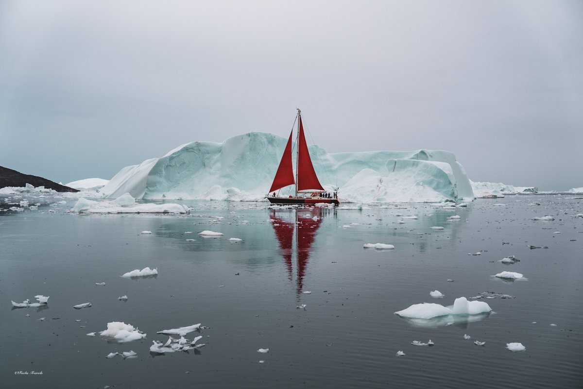 Red sailboat dwarfed by a towering white iceberg in the calm waters of Disko Bay at sunset