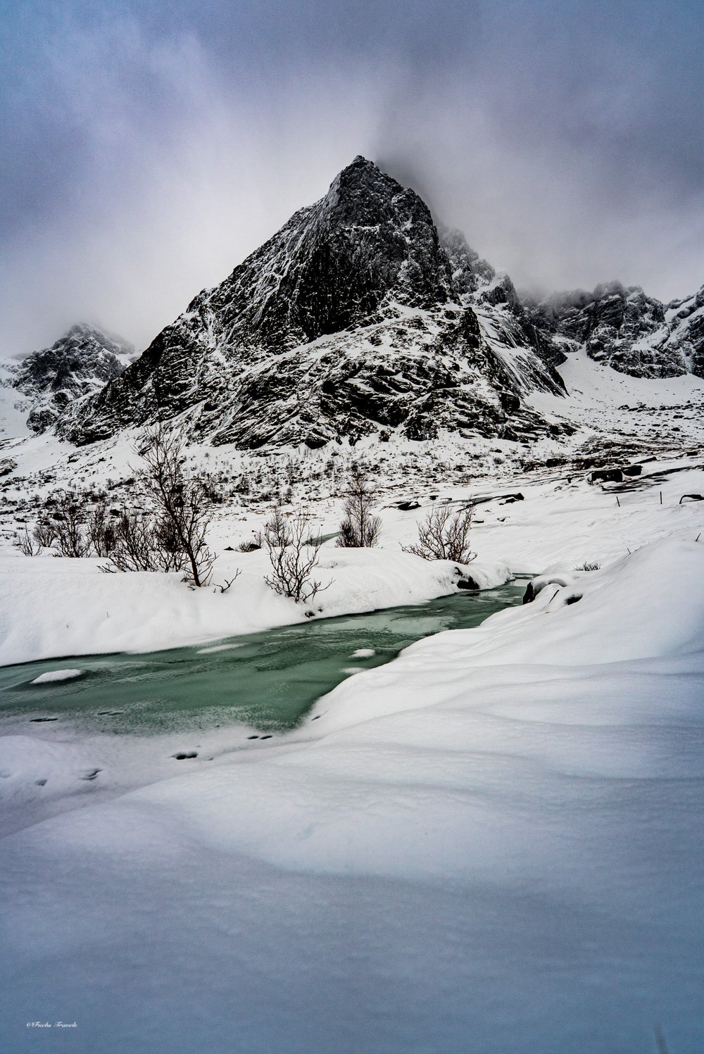 Dramatic pyramidal peak rising above a turquoise frozen stream in the Lofoten winter landscape